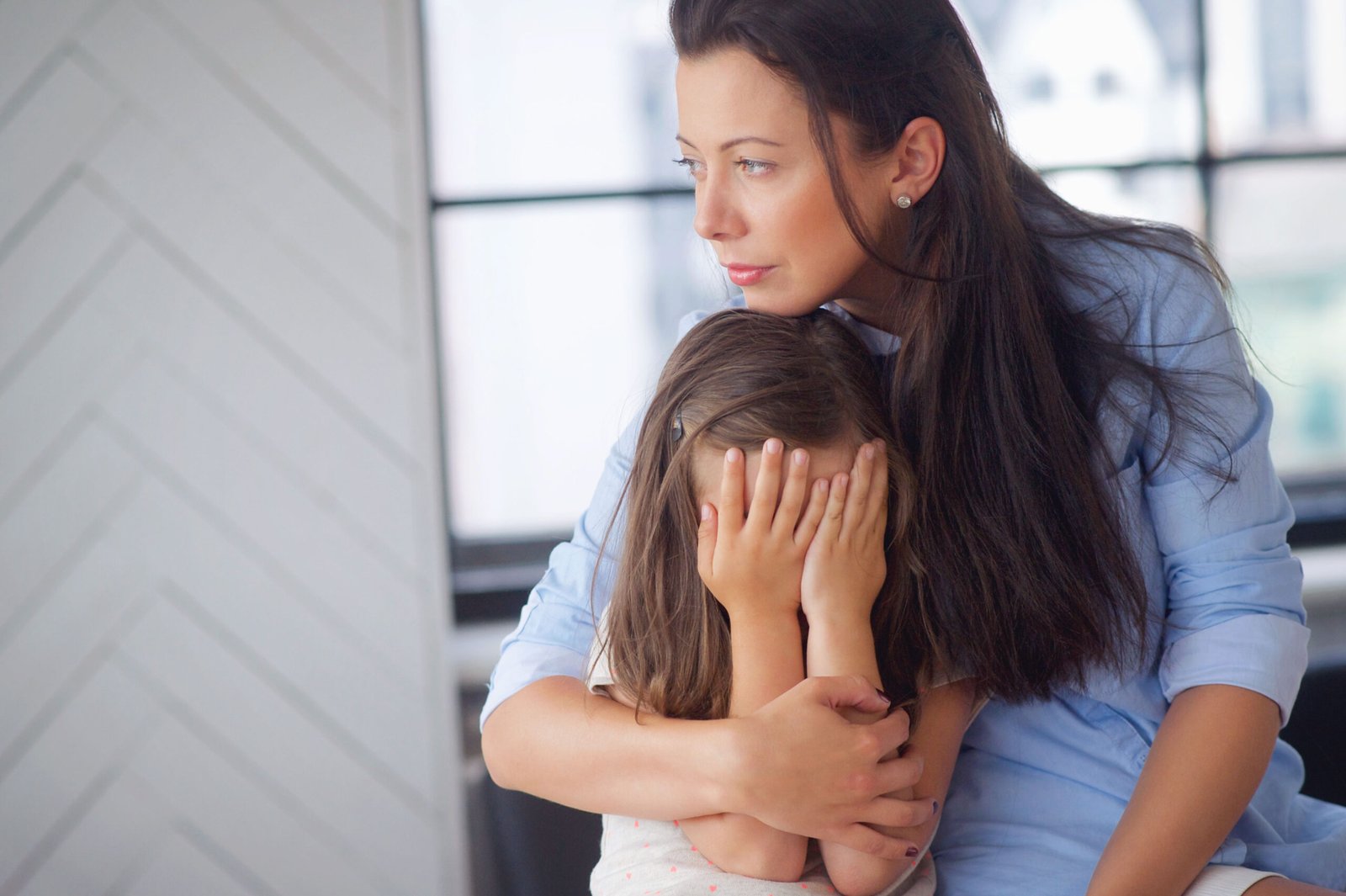 Mother gently caressing and calming her crying baby son while standing in a living room, providing comfort and care.