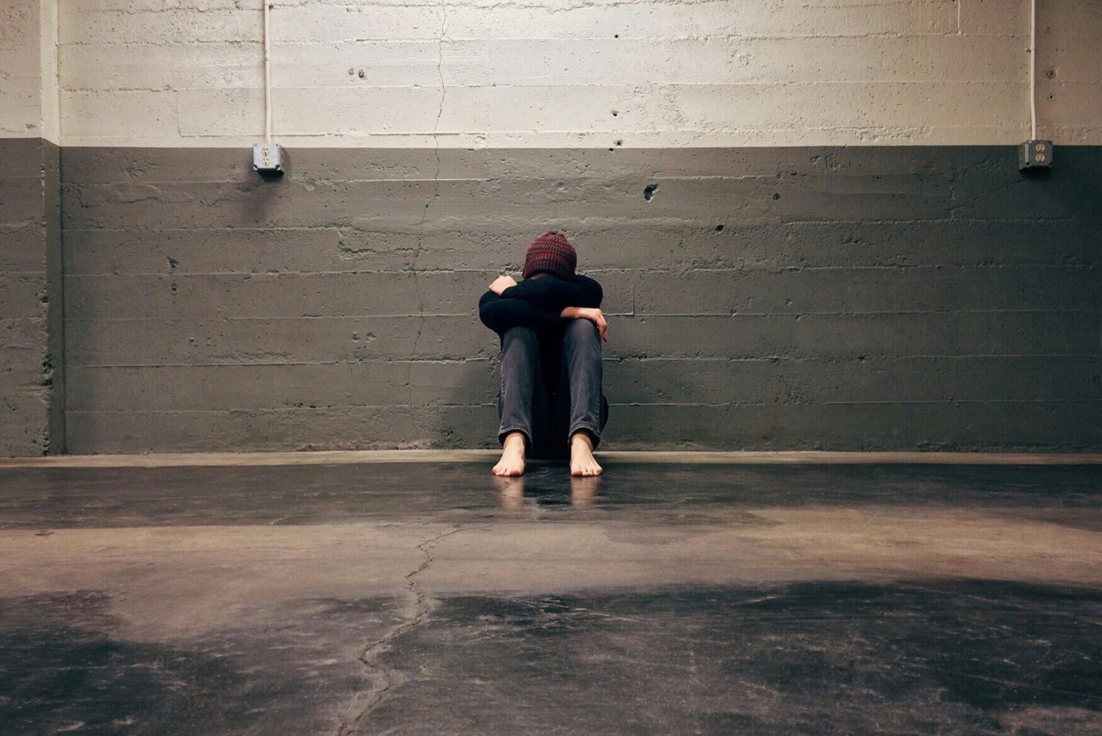 Person sitting against a wall with head down, leaning on knees
