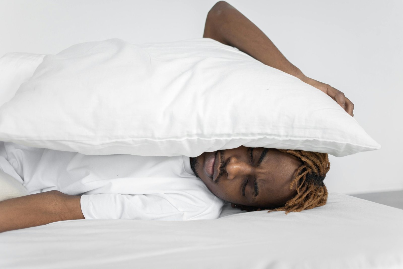 Man in bed with a pillow over his head, frustrated that he can’t fall asleep